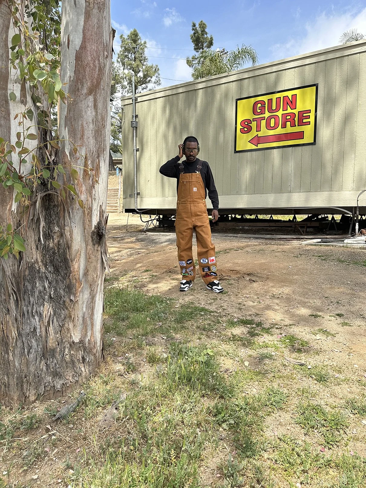 Clarence Nathaniel, posing outdoors near a tree, talking on his cellphone, wearing brown work overalls with patches, black long-sleeve shirt, and sneakers. Behind him is a mobile building with a yellow sign that says 'GUN STORE'.