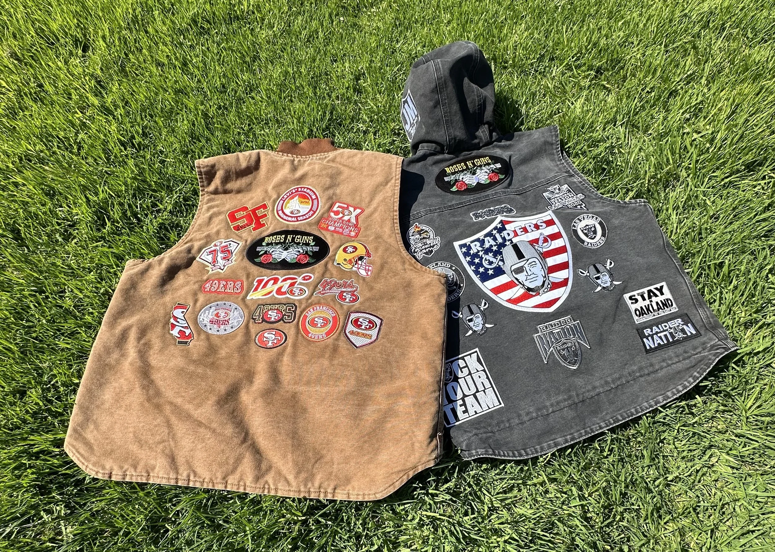 Two vests, one tan and one black, covered with various San Francisco 49ers and Raiders patches, lying on green grass.