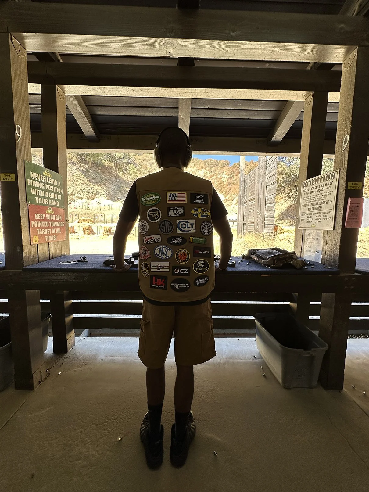 A person at an outdoor shooting range, standing in a shooting booth, wearing headphones and a vest covered with gun brand patches, aiming at the target.