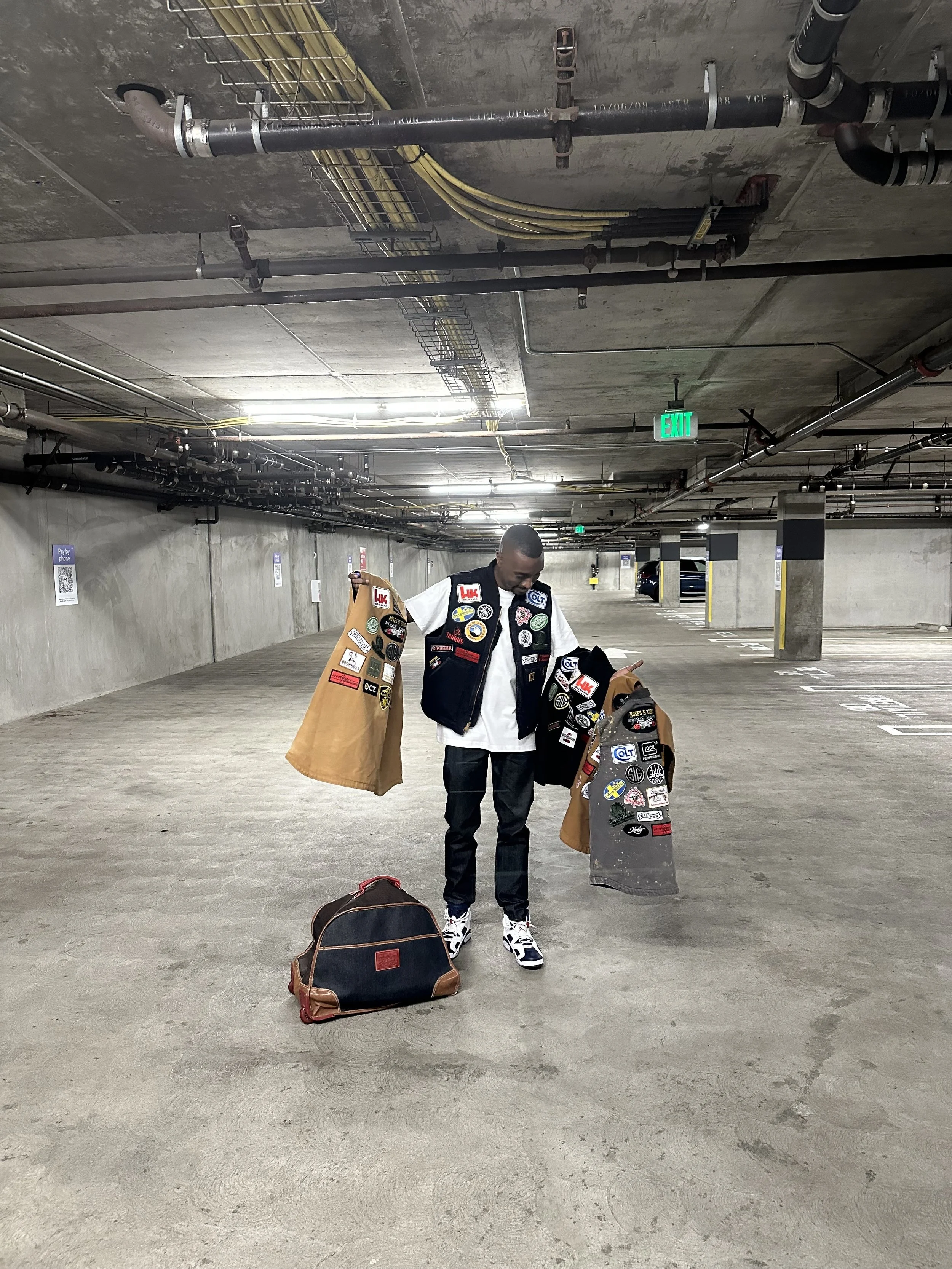 Founder of Roses N' Guns, Clarence Nathaniel, is standing in an parking garage holding two a bunch of vests decorated with embroidered patches, and has a black bag at his feet. The garage has concrete walls and ceiling, with pipelines.