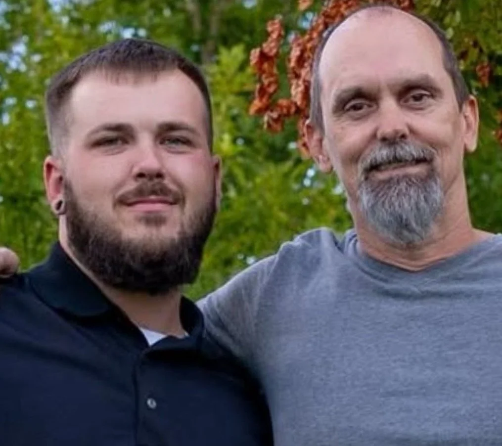 Two men standing outdoors in front of green trees, smiling at the camera. One man has a beard and earrings, the other has a mustache and goatee.