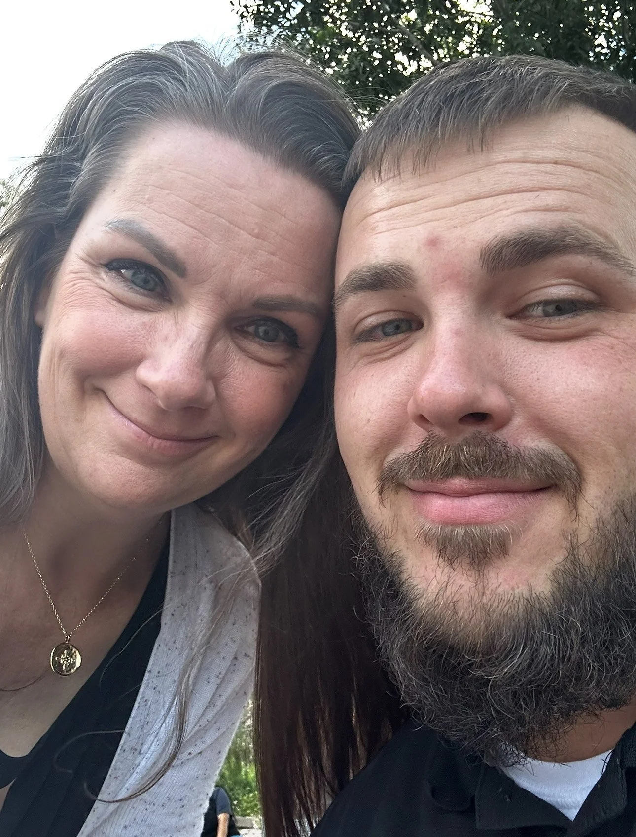 Close-up selfie of a smiling woman with blue eyes and long brown hair, and a smiling man with a beard and mustache, outdoors with trees in the background.