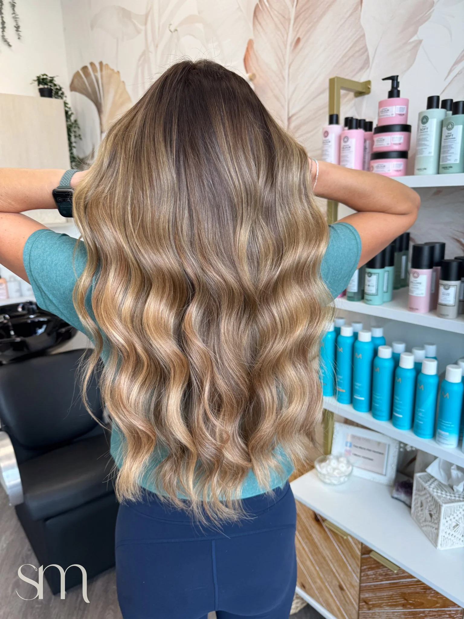 Back of a woman with long, wavy, blonde hair in a salon, standing in front of shelves with hair care products.