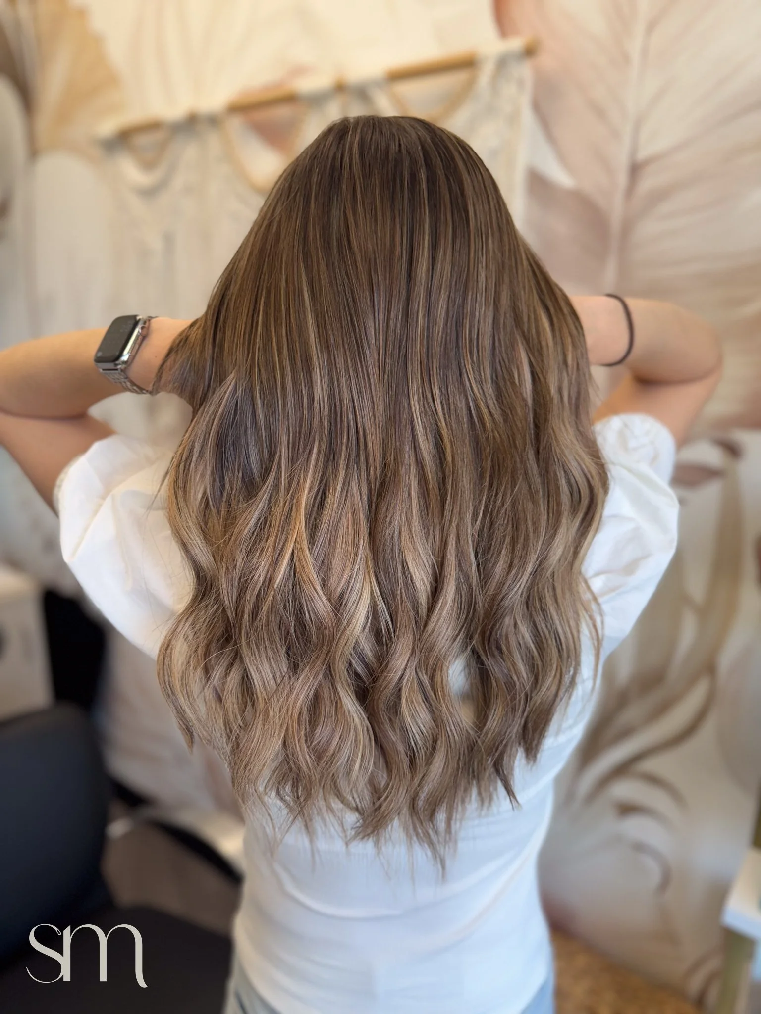 Back view of a woman with long wavy light brown hair wearing a white shirt, with arms raised behind her head, standing indoors.