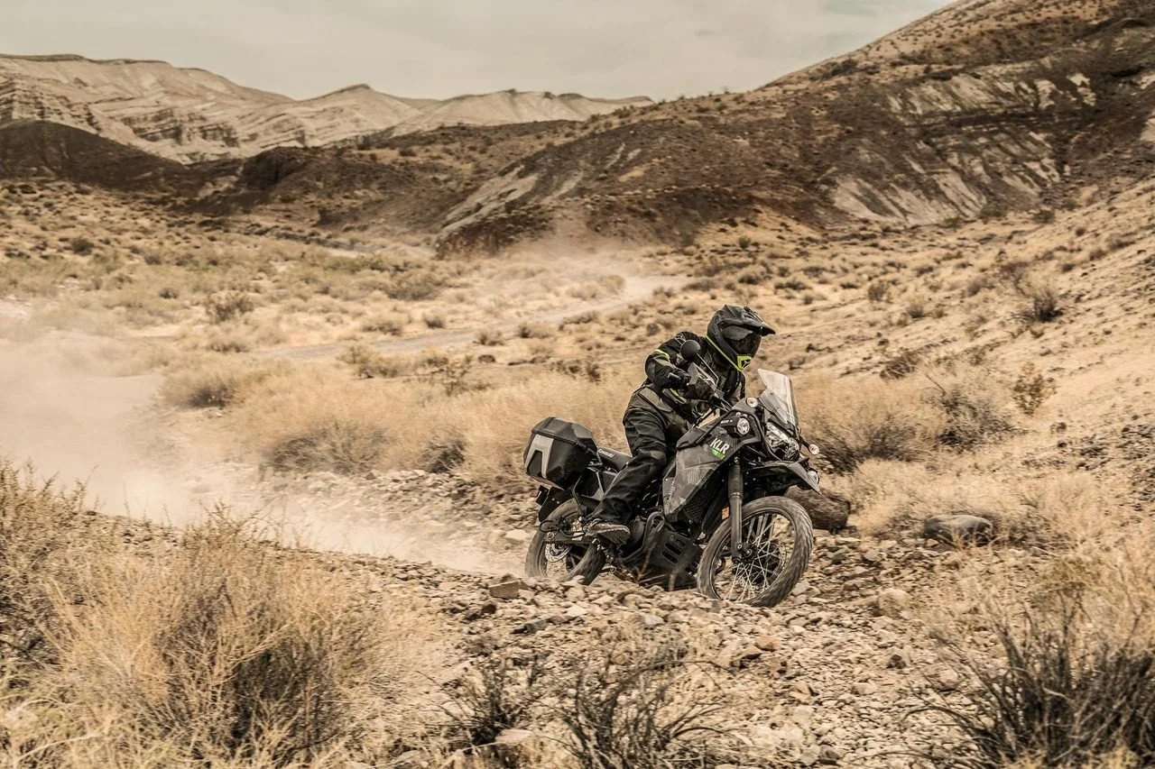 Motorcyclist wearing black gear and helmet riding a dirt bike through a dry desert landscape with mountains in the background.