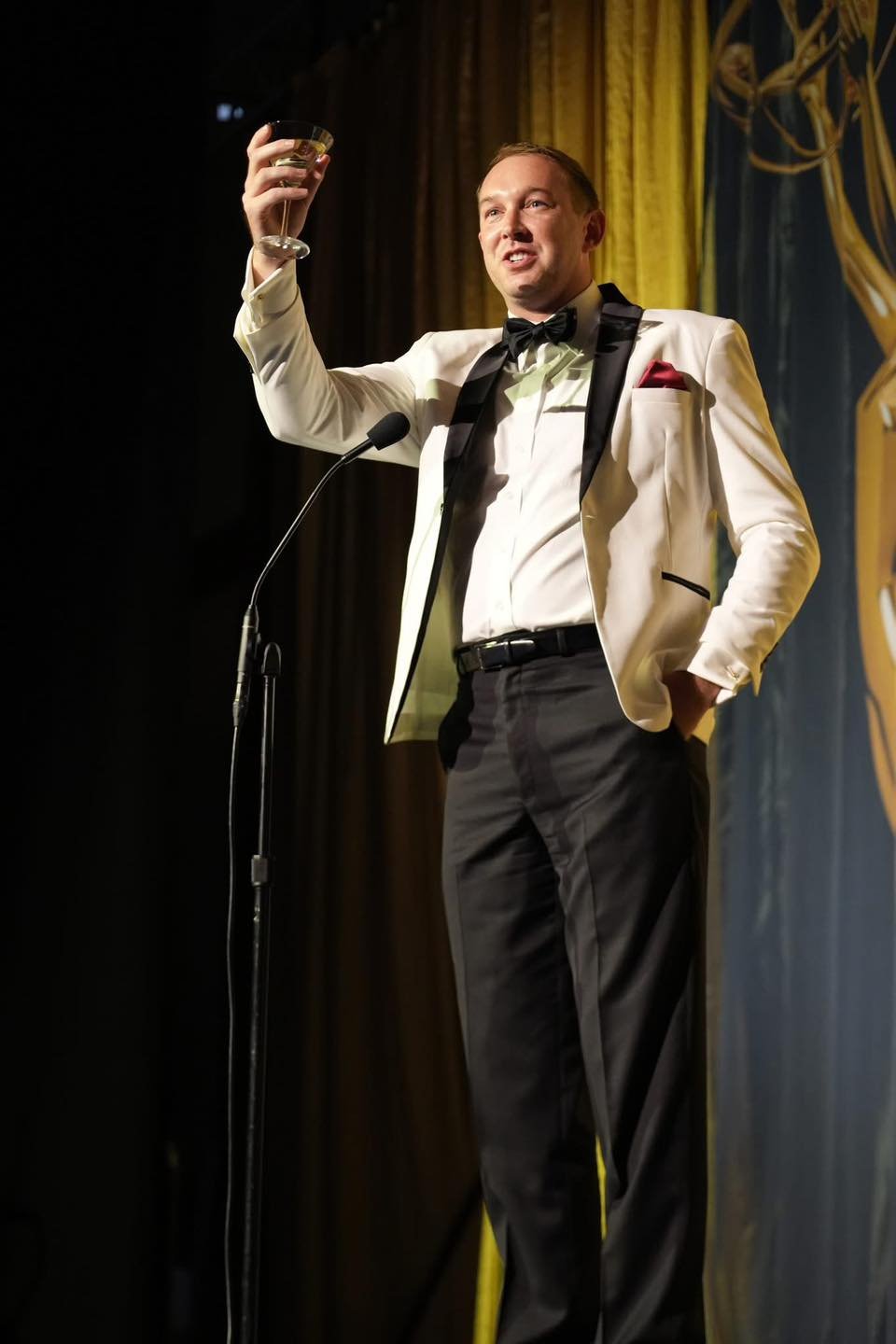 EVan wears a white tuxedo jacket while lifting a glass to cheers an audience