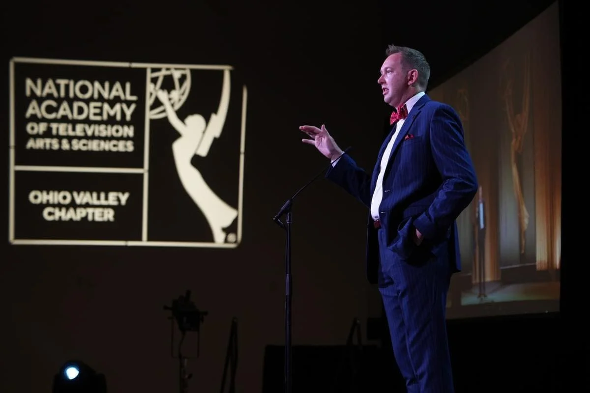 the National Academy of Television Arts and Sciences logo is projected on a wall next to Evan as he speaks and wears a blue suit and bowtie