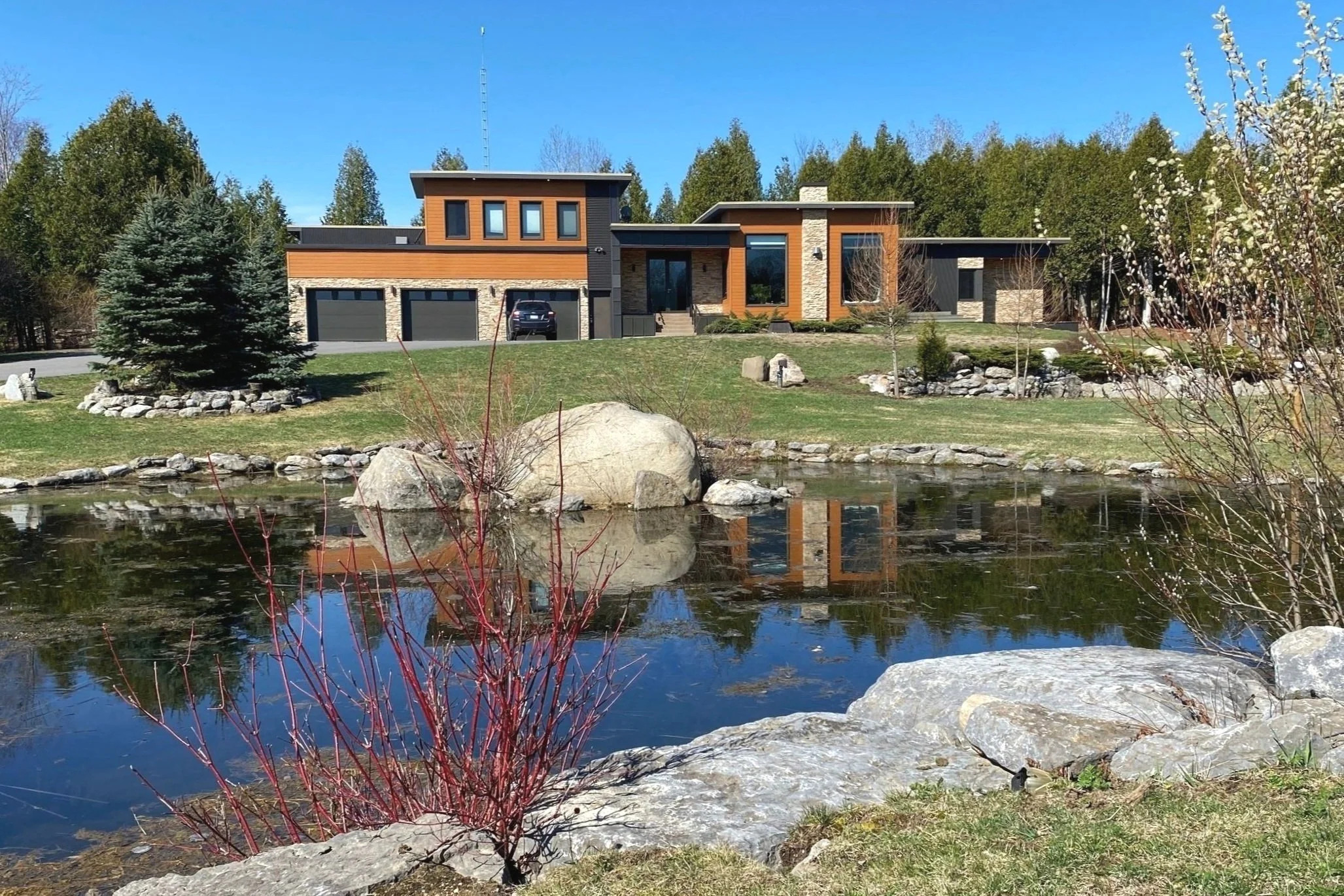 Modern house with large windows and a flat roof, surrounded by trees and landscape. A pond with rocks is in the foreground, reflecting the house and sky.