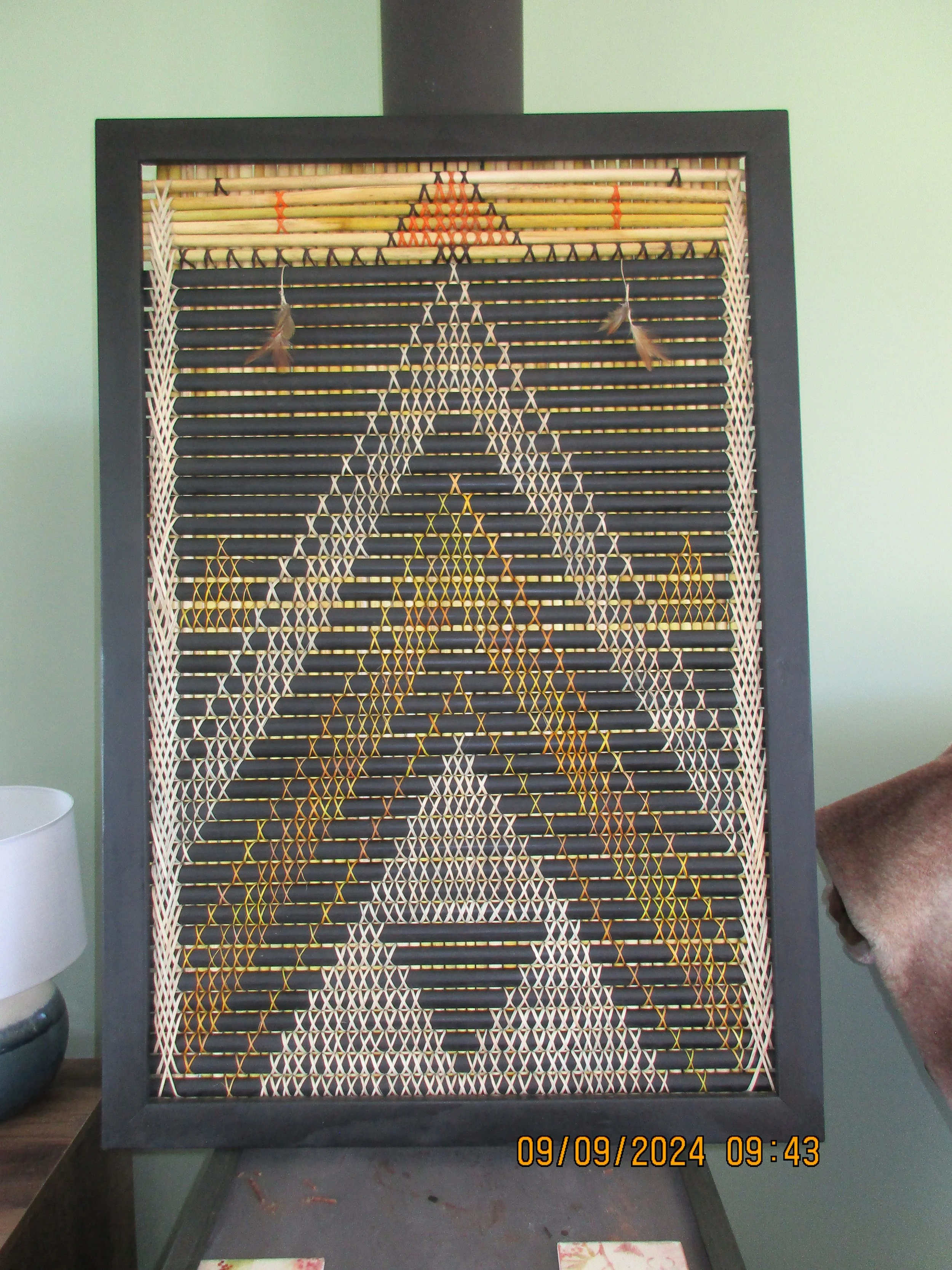 A woven bamboo or rattan patterned decorative piece framed in black, decorated with small feathers hanging from strings.