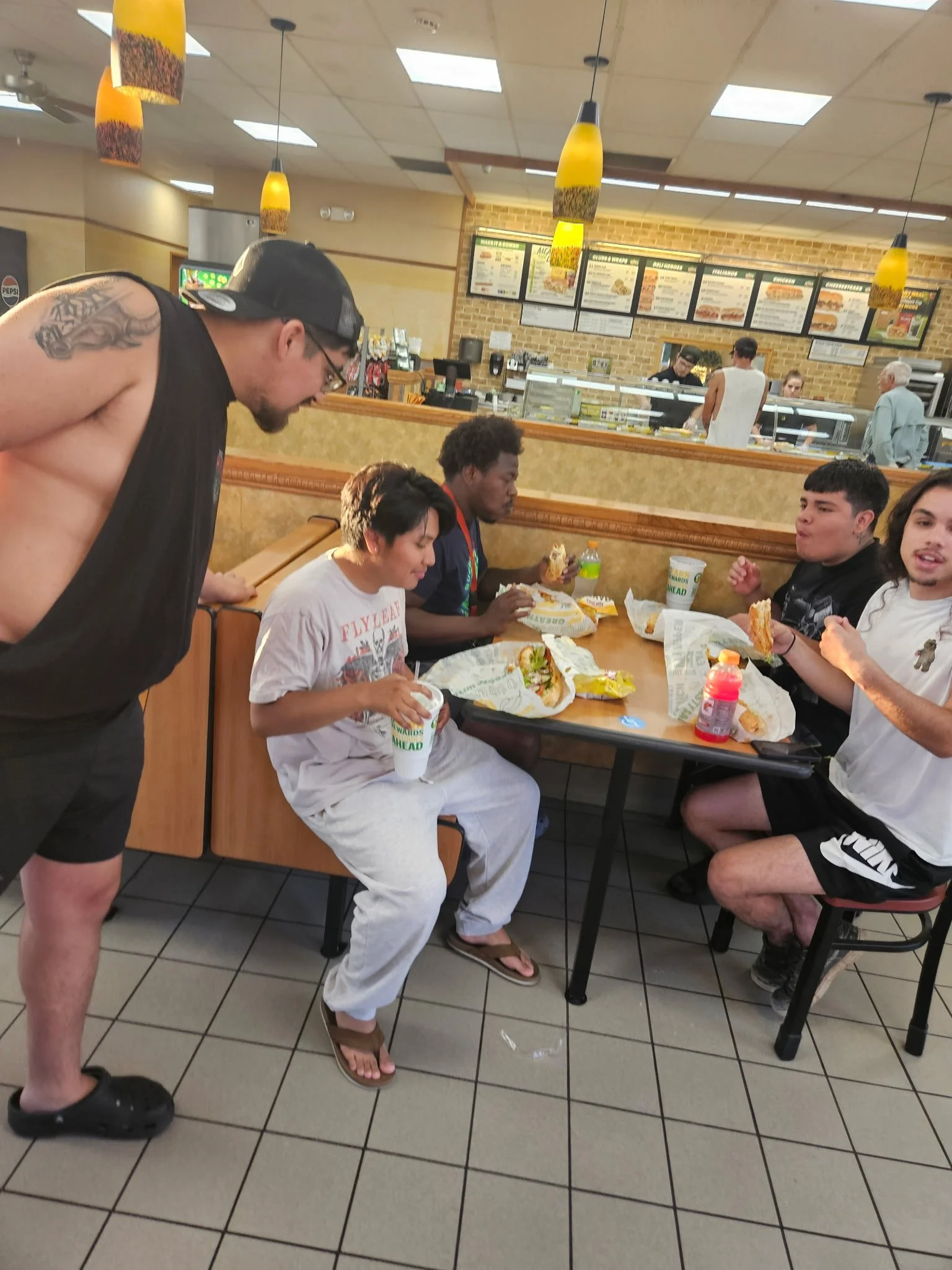 A group of five young people sitting at a table in a fast food restaurant, eating sandwiches. One person is standing, leaning over the table. The restaurant has a menu display on the brick wall and yellow pendant lights hanging from the ceiling.