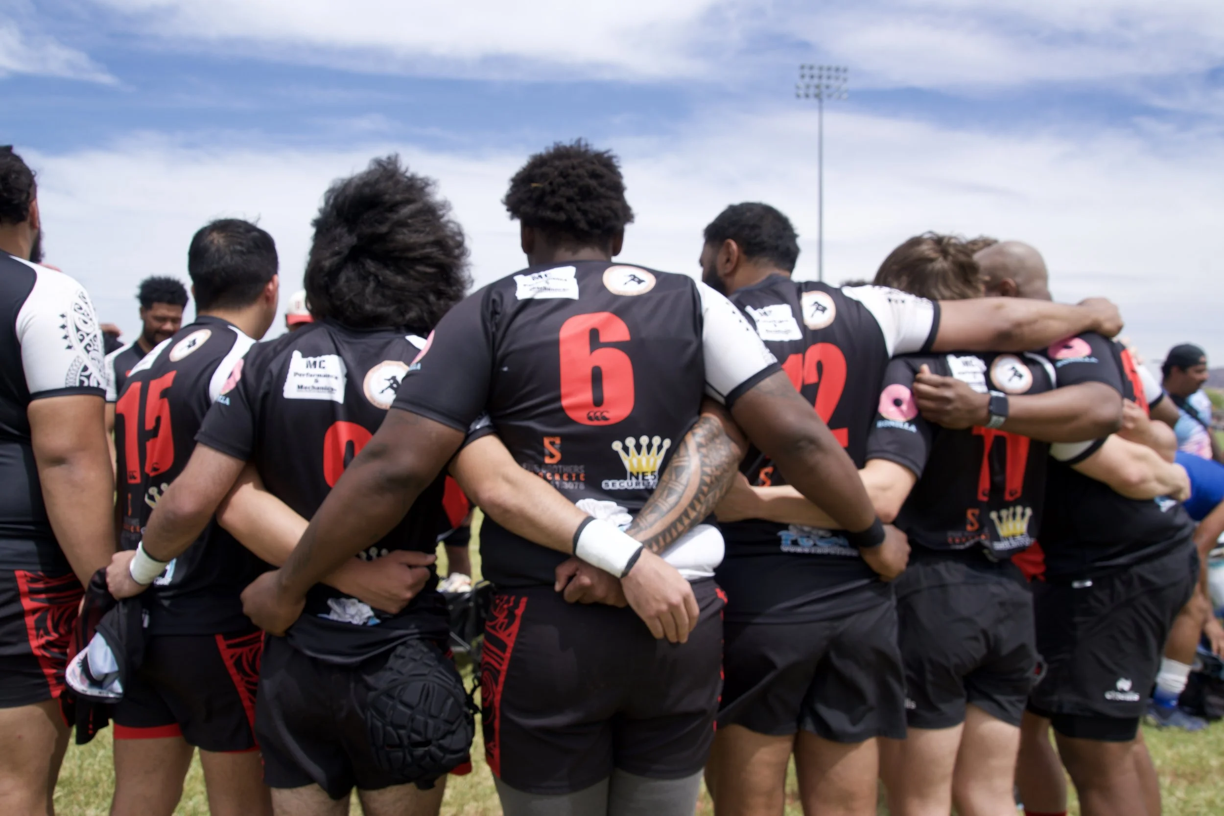 Rugby team huddled together on the field during a game, some with arms around each other's shoulders, under a partly cloudy sky.