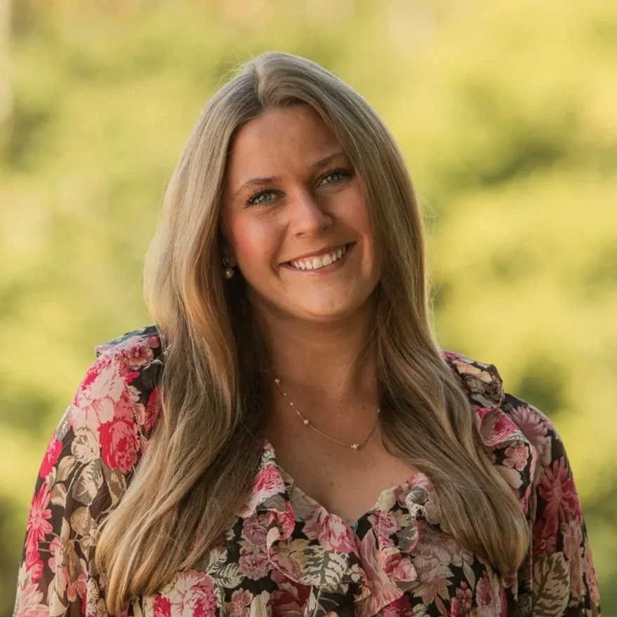 A woman with long wavy blonde hair, wearing a brown sleeveless dress and a cross necklace, smiling outdoors with a blurred background of trees and a structure.