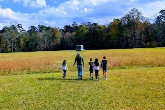 A family walking in a grassy field, holding hands, with children and an adult woman in a red floral dress and straw hat in the center, a man in a denim jacket carrying a baby girl on the right, and two boys in front, one pulling on a rope and the other smiling and holding the mother's hand, during sunset with string lights in the background.