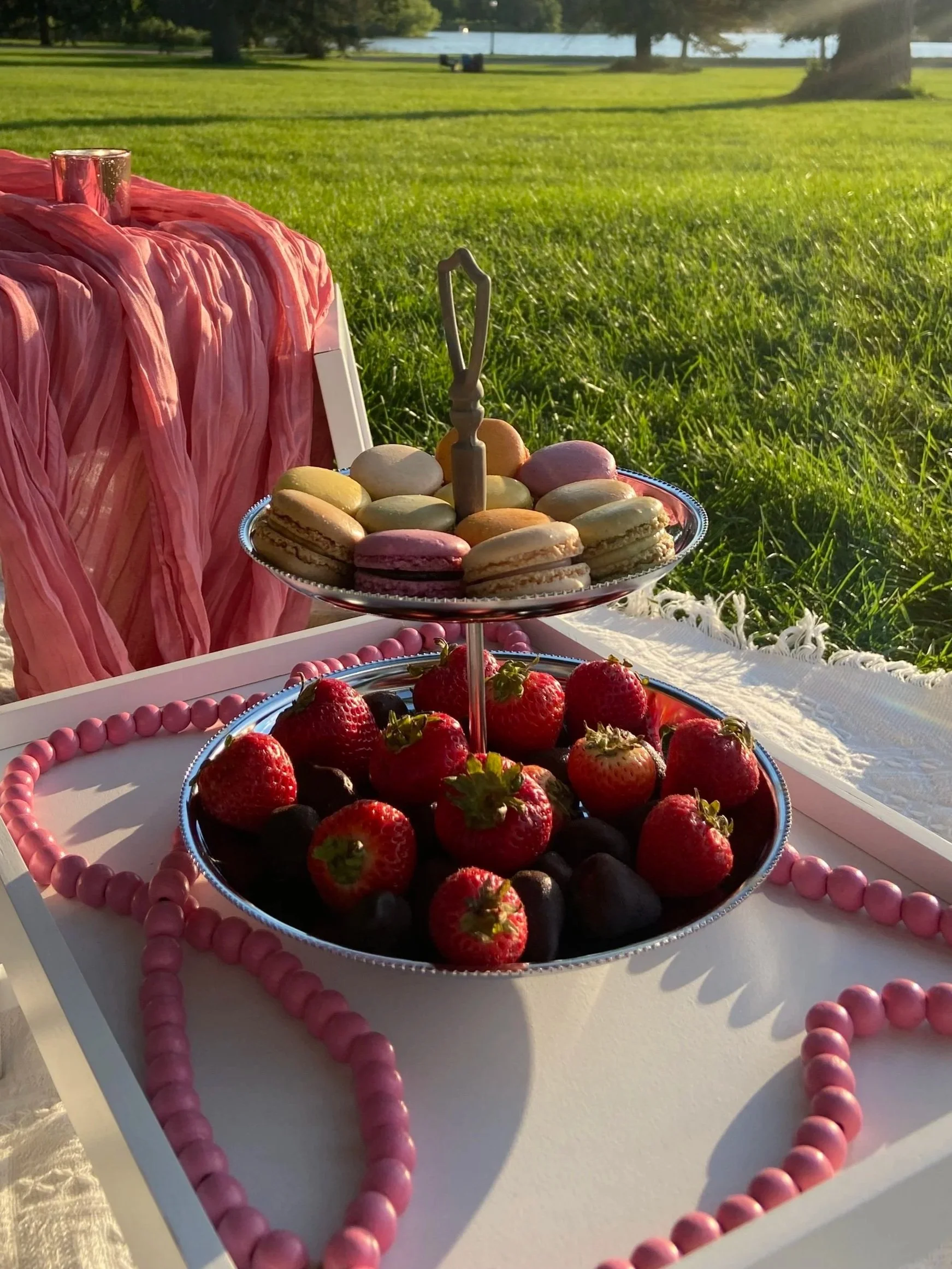 A two-tiered tray with colorful macarons on the top tier and sprinkle-covered Rice Krispies treats on the bottom. Nearby, a plate with assorted mini cupcakes with colorful icing and sprinkles. The display is set on a white tablecloth with a decorative floral background.