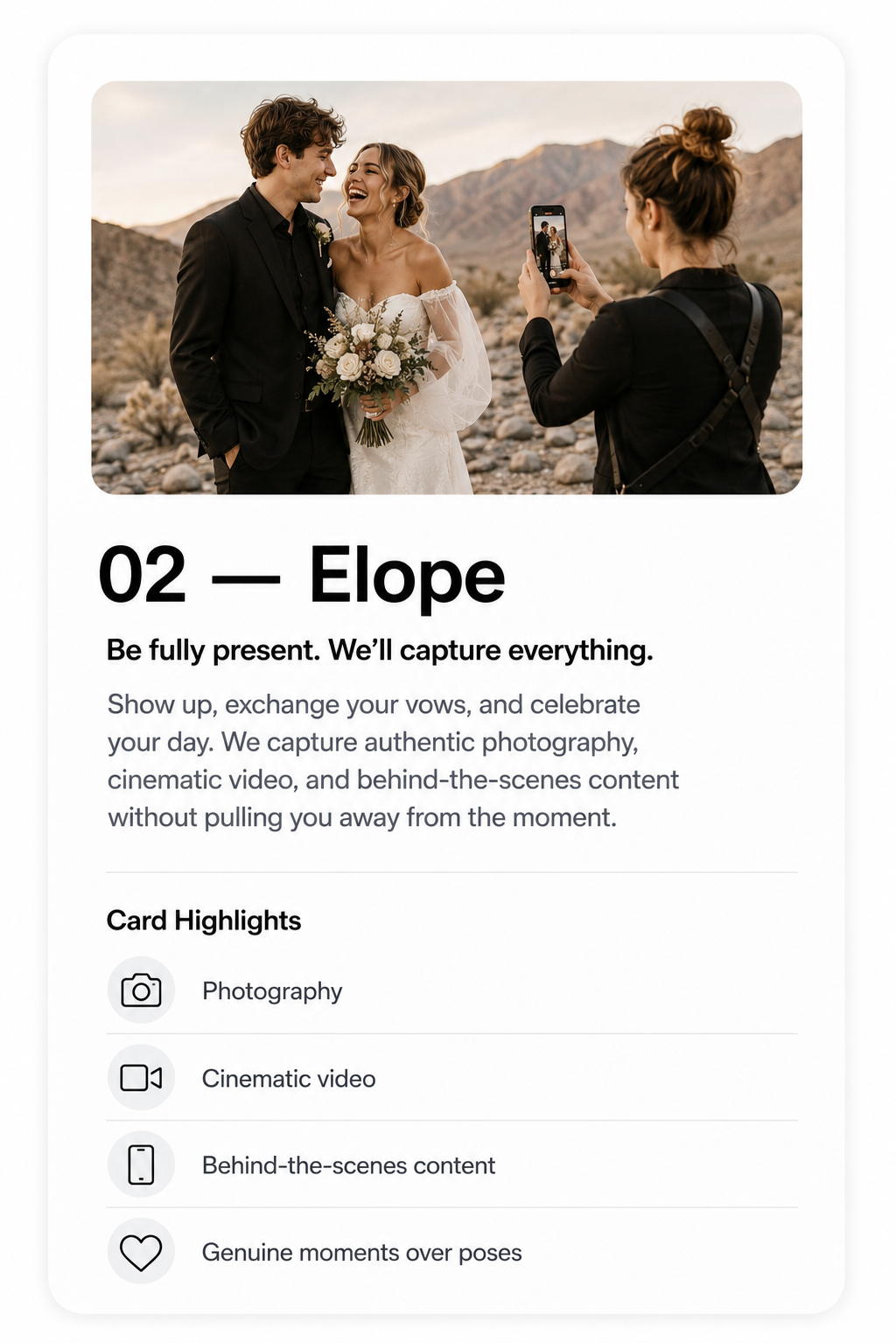 A couple dressed in wedding attire standing in a desert landscape, smiling and looking at each other while a person takes their photo.