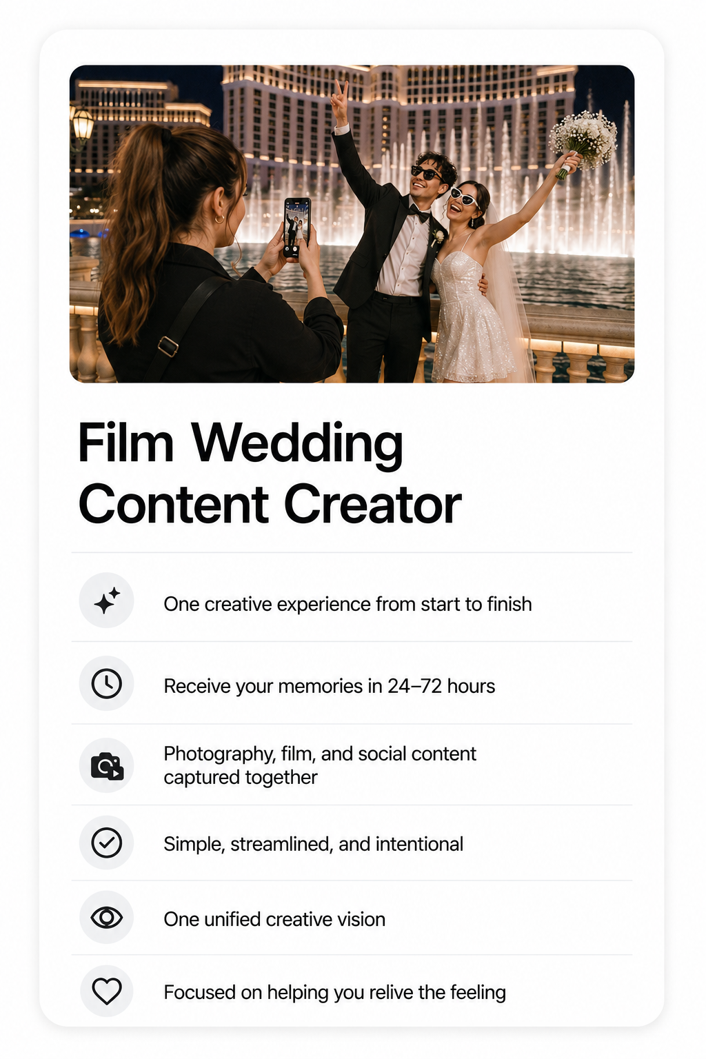 A wedding couple taking a photo at night with a fountain and hotel behind them, the groom in a black tuxedo and the bride holding a bouquet, as another person captures the moment with a smartphone.