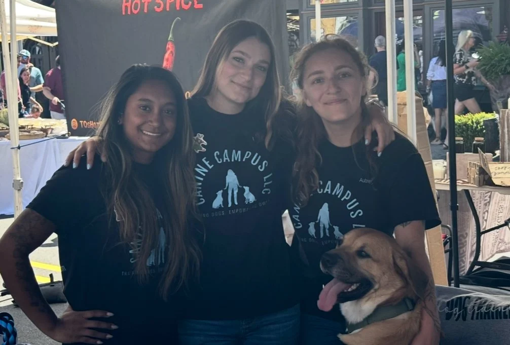 Three women standing close together at an outdoor event, smiling, with a large dog, possibly a Labrador Retriever, sitting in front of them. They are wearing black t-shirts with a logo and text related to a canine campus.