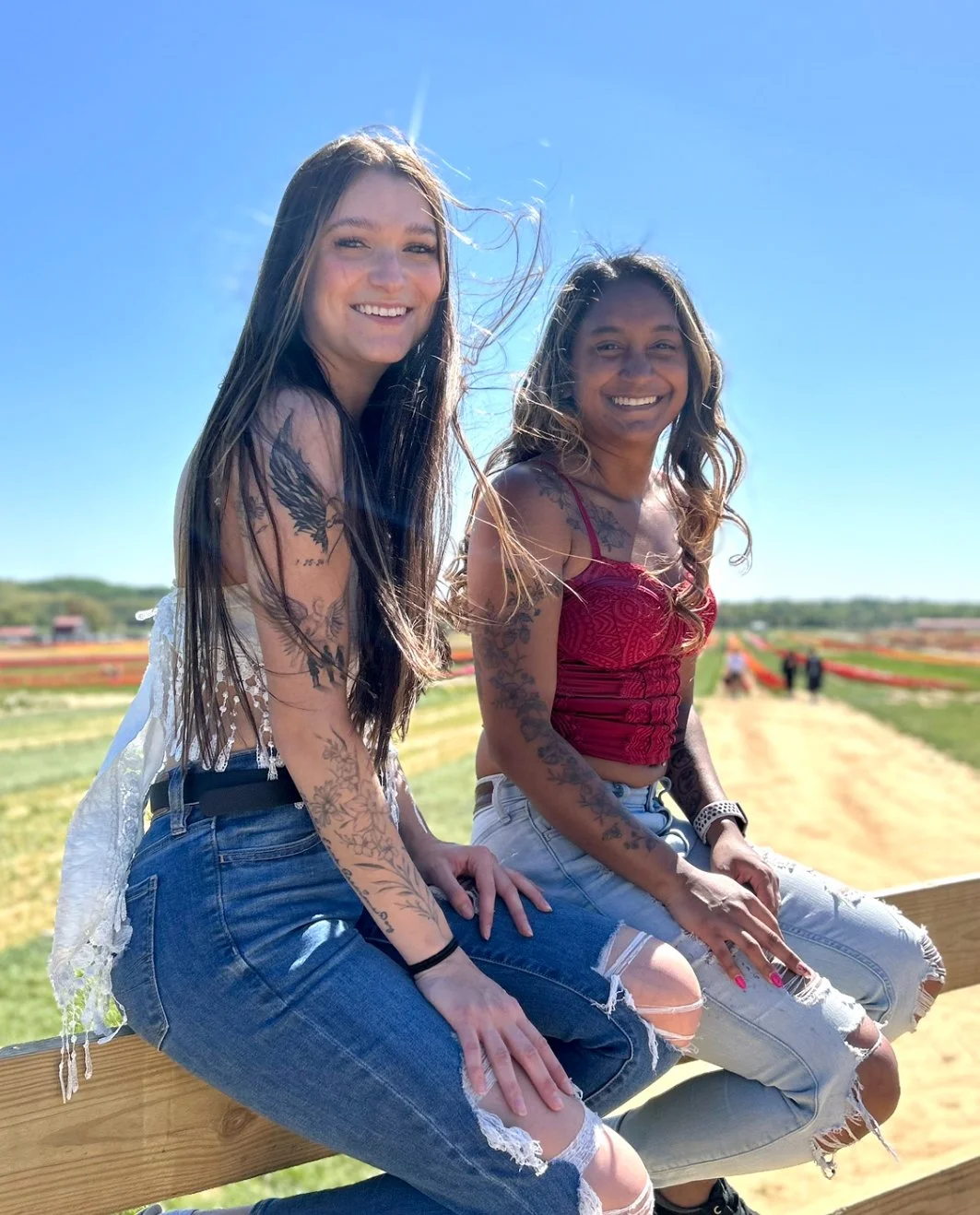 Two young women with tattoos sitting on a wooden fence outdoors under a clear blue sky, smiling at the camera, with a field and pathway in the background.