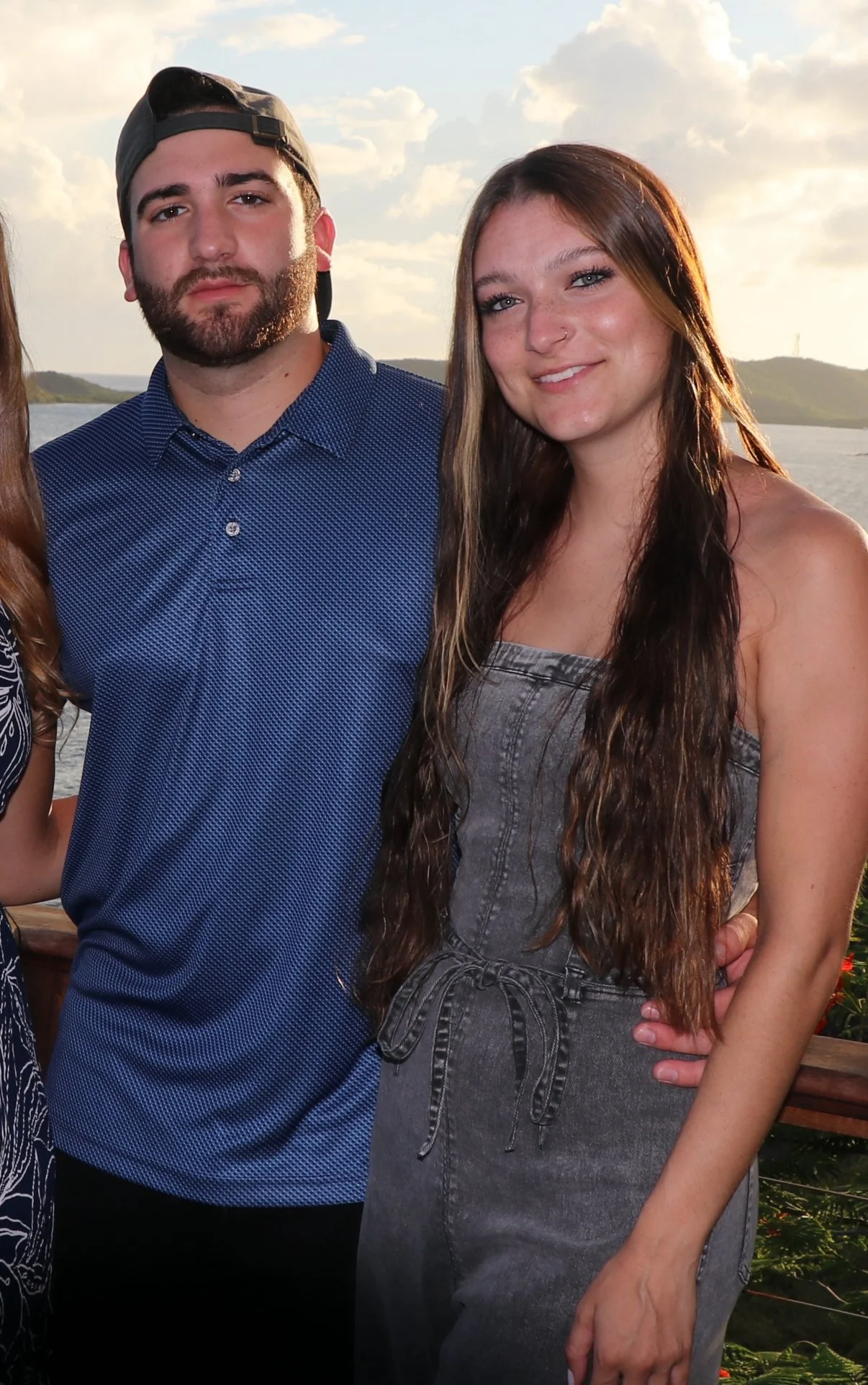 A young woman with long brown hair, wearing a gray strapless dress, standing next to a young man with a beard, wearing a blue button-up shirt and a backward gray cap, outdoors near a body of water during sunset.