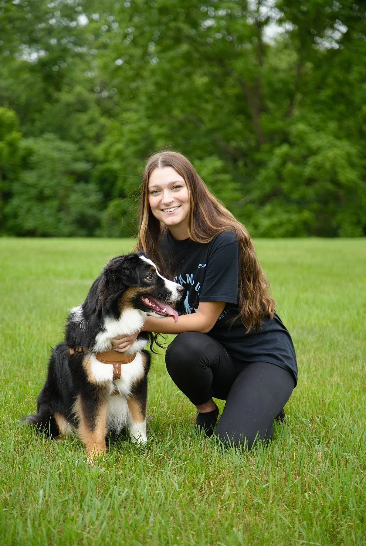 A young woman kneels on the grass, smiling, with a black, white, and tan Australian Shepherd puppy sitting beside her outdoors in a lush green park.