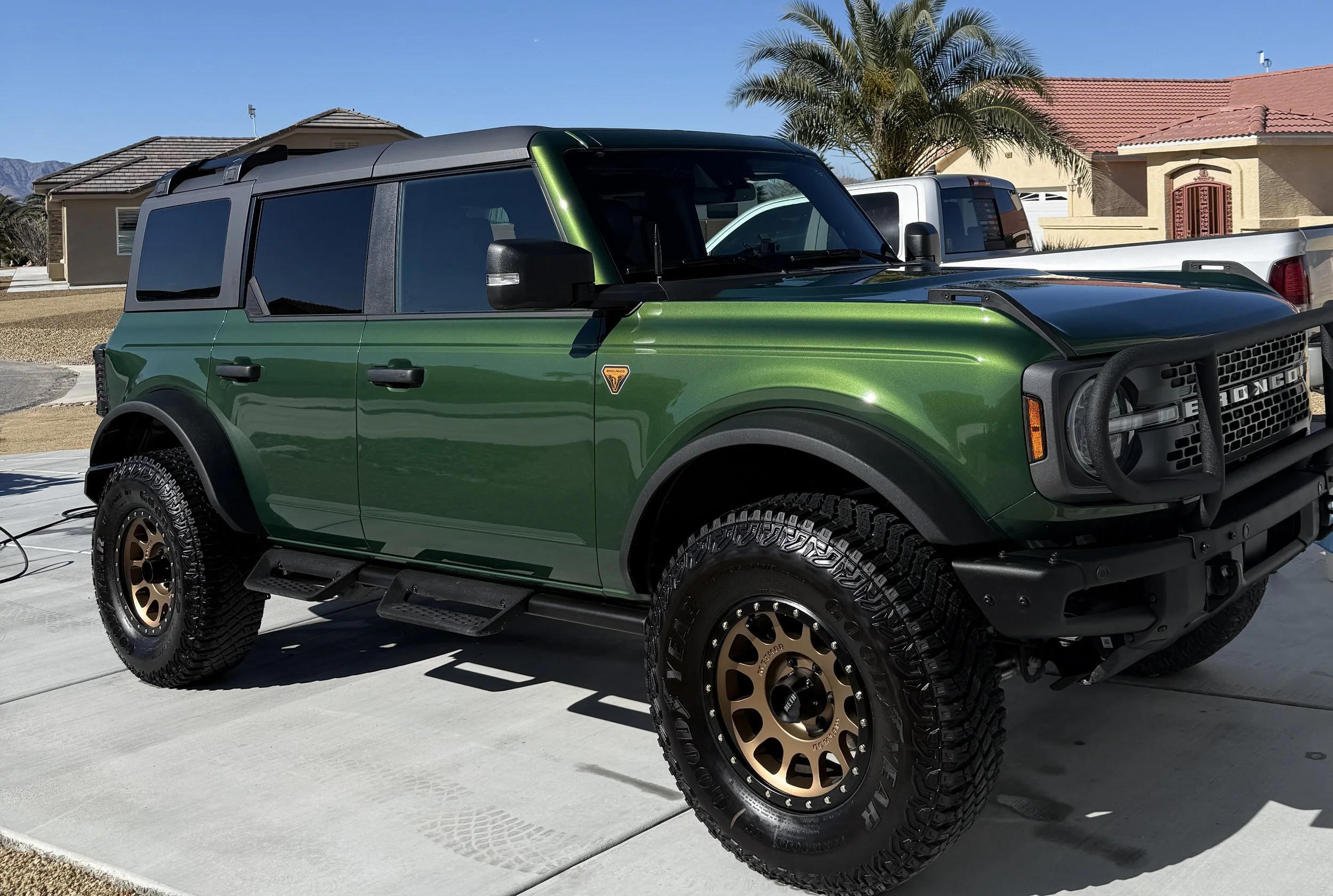 A green off-road SUV with large tires and black bumper, parked in a driveway with a house and palm tree in the background.