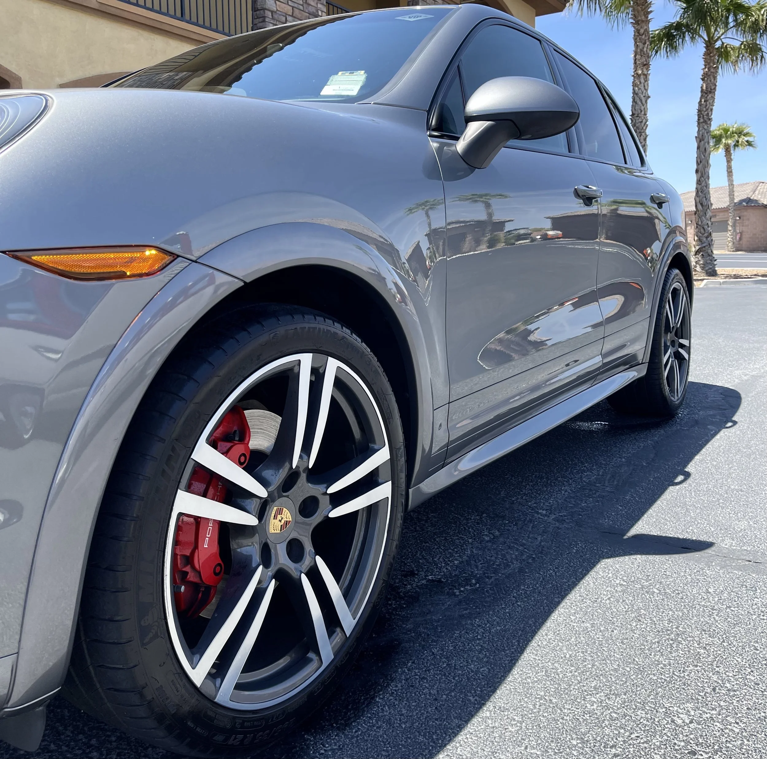 Side view of a gray Porsche SUV with black alloy wheels and red brake calipers, parked on a sunny street with palm trees in the background.