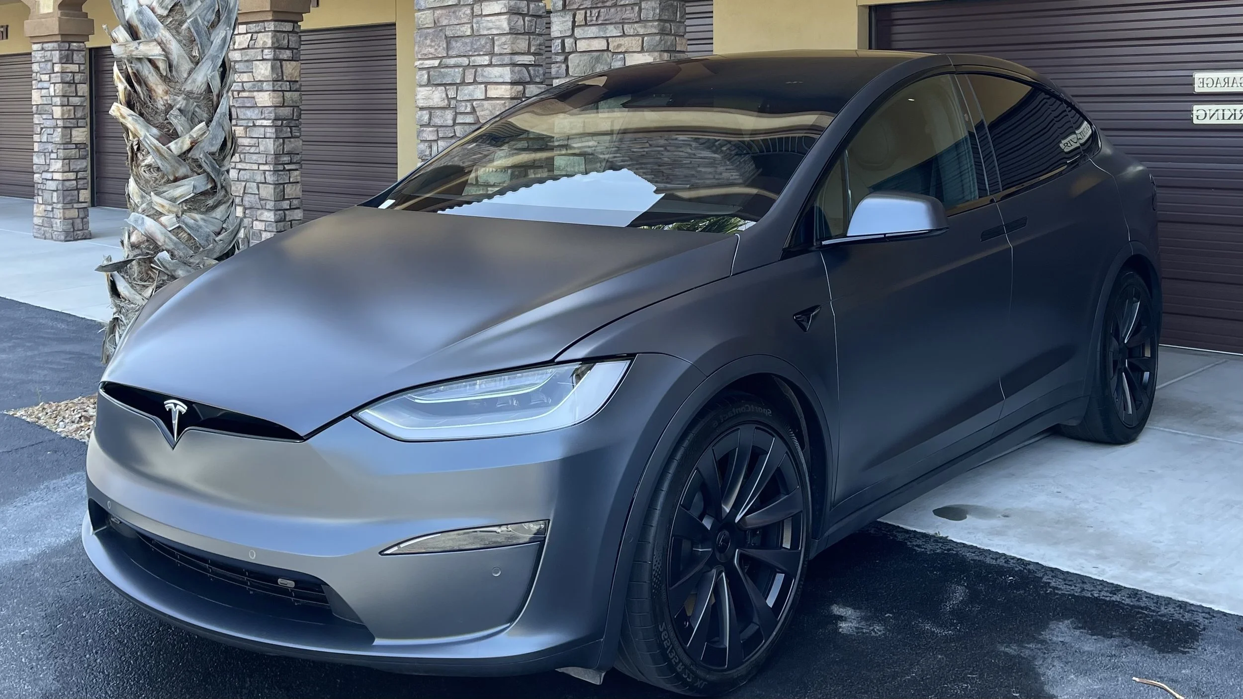 A matte black Tesla Model Y parked in front of a garage with stone and yellow siding exterior, next to a palm tree.