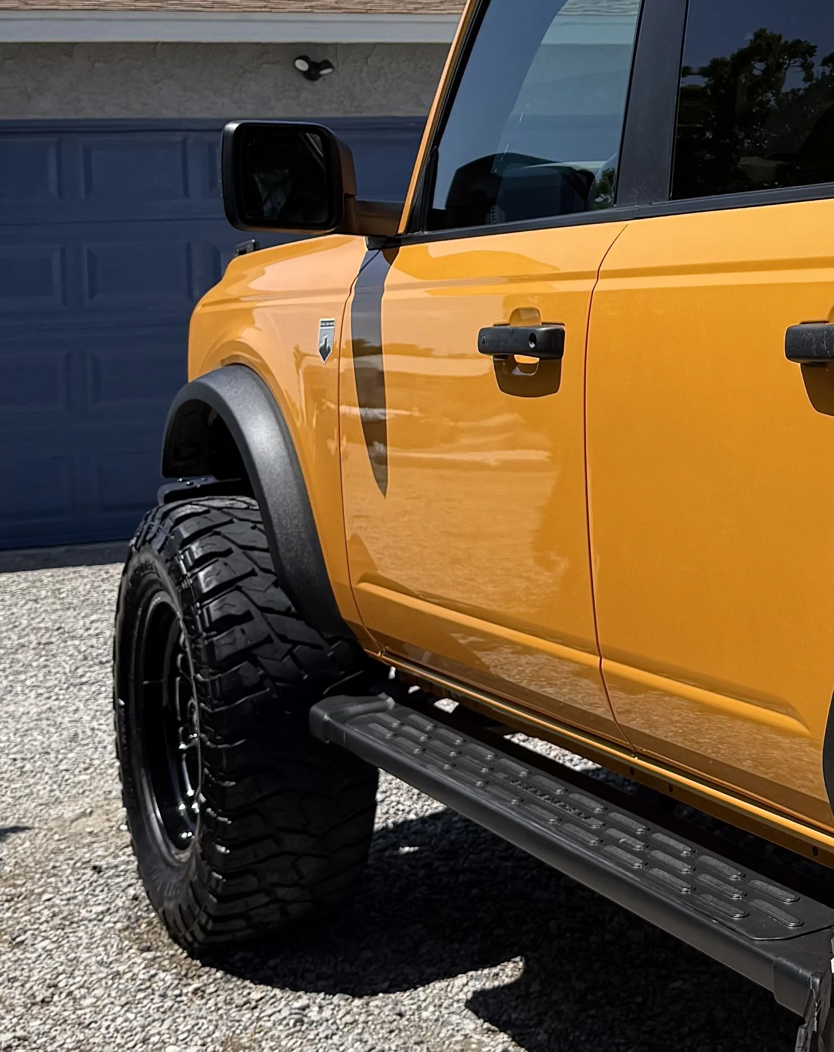 Close-up of a yellow off-road vehicle showing the front side, including large tire, side mirror, door handle, and running board, parked in front of a blue garage door.