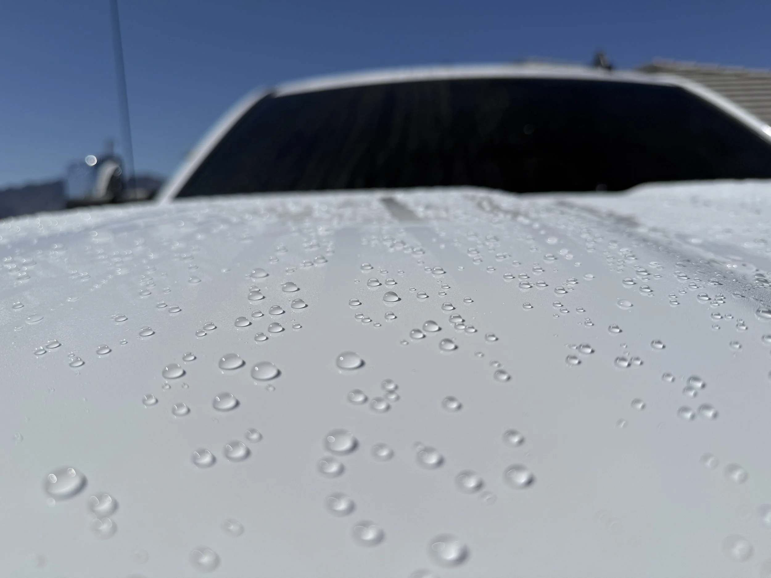 Close-up of water droplets on a white surface with a blurred vehicle in the background under a clear sky.
