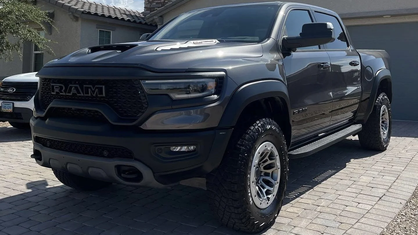 A black Ram pickup truck parked on a brick driveway with a house and another vehicle visible in the background.