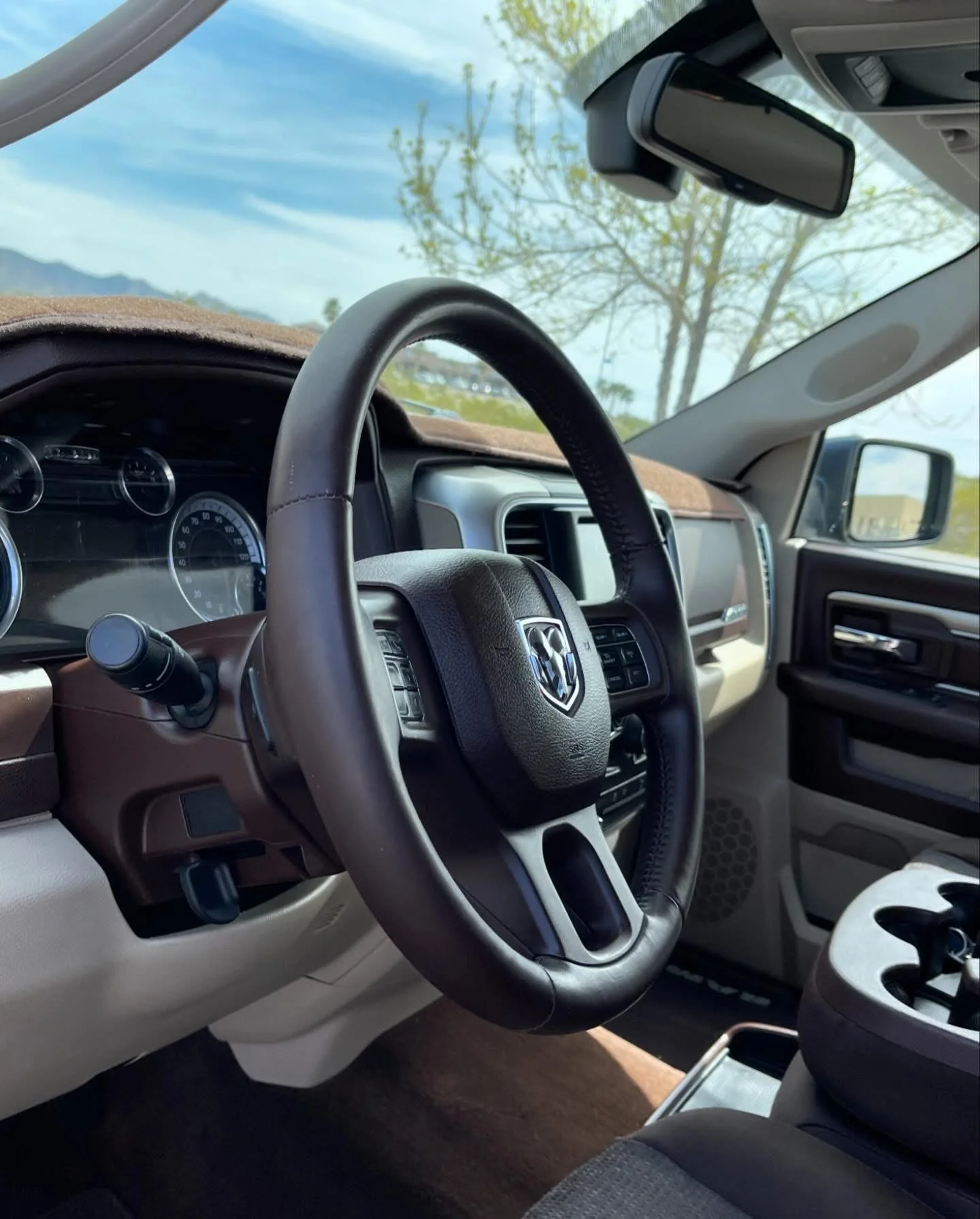 Interior of a truck with a steering wheel featuring the Dodge logo, dashboard, and a view of trees and sky through the windshield.