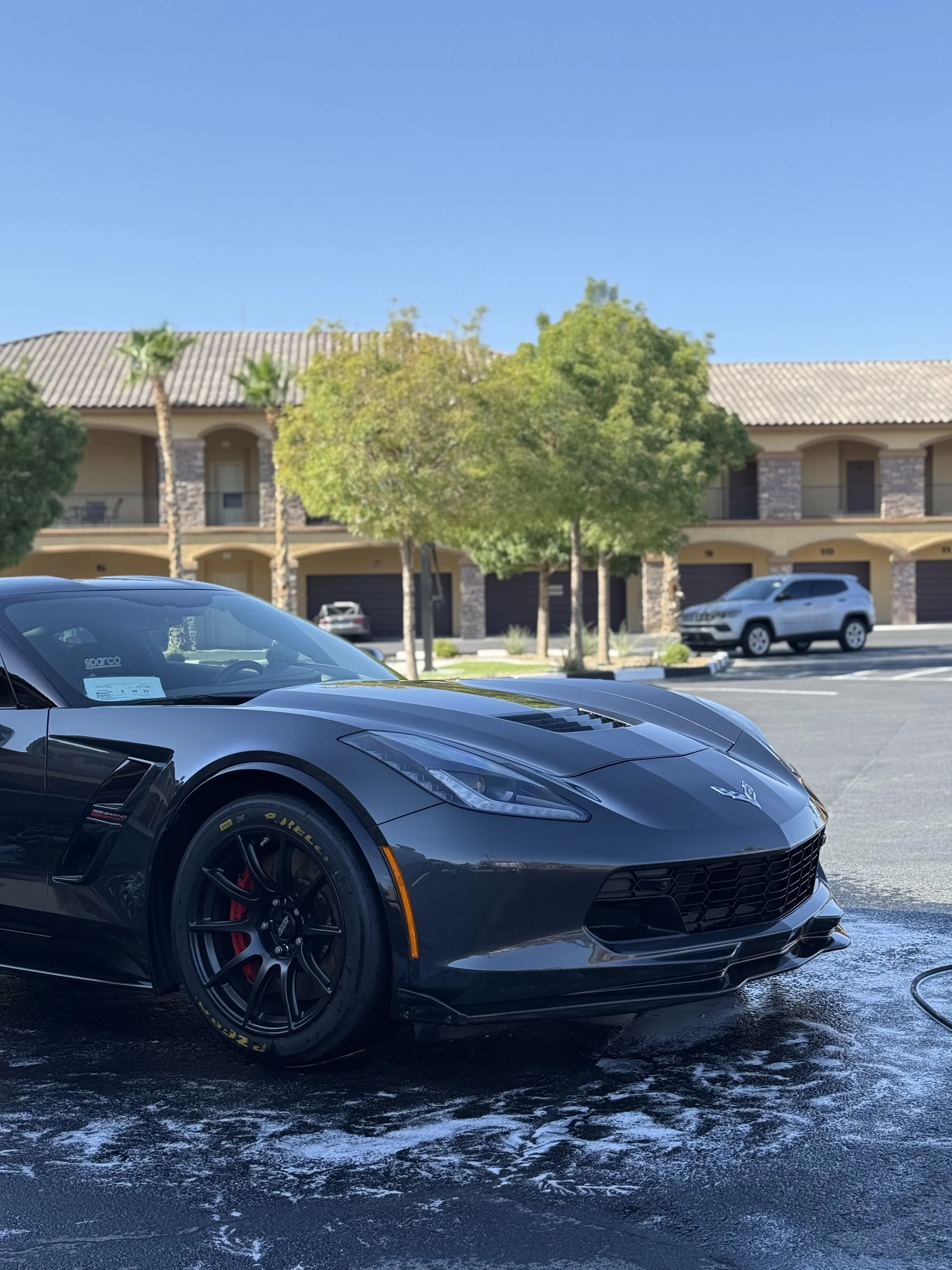 Black Chevrolet Corvette parked on a car wash station with soap and water on the ground, residential buildings with balconies, trees, and a clear blue sky in the background.