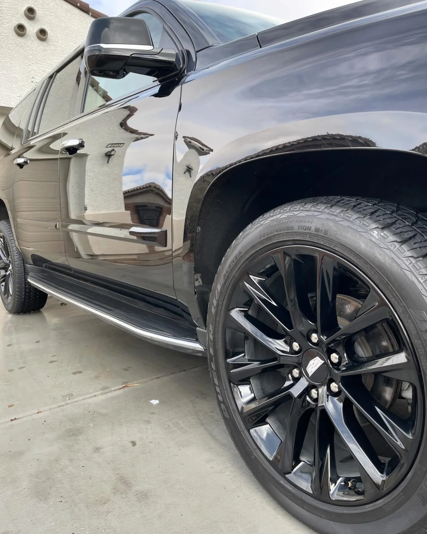 Close-up of a black luxury SUV showing the front wheel and part of the side, with reflections of the sky and nearby building on the shiny surface.