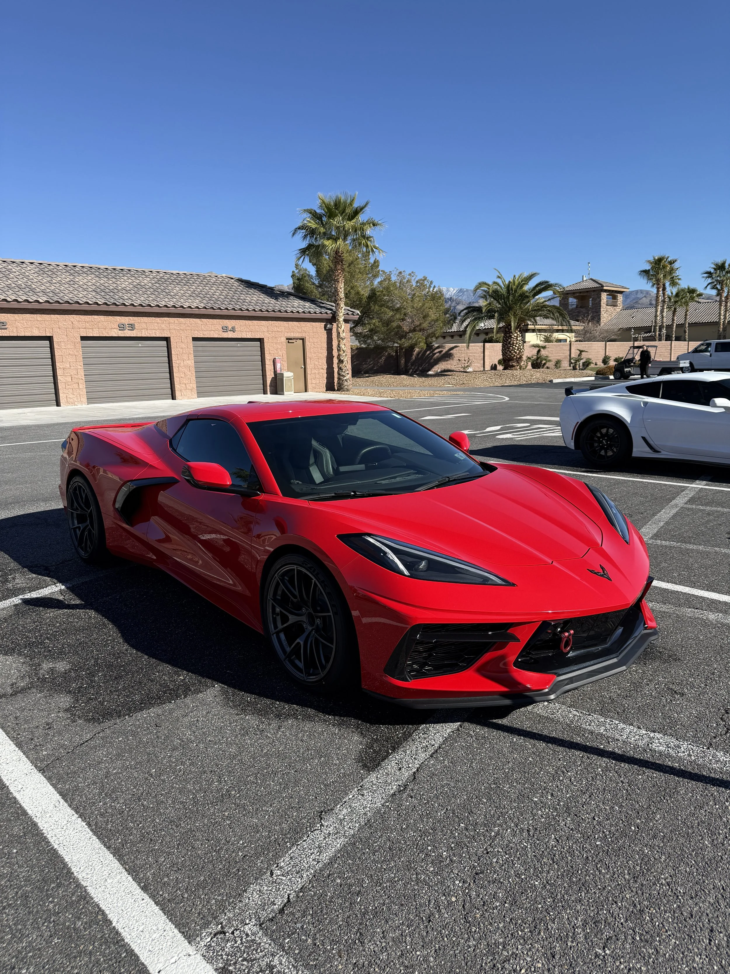 A red sports car parked in a parking lot with palm trees and a clear blue sky in the background.