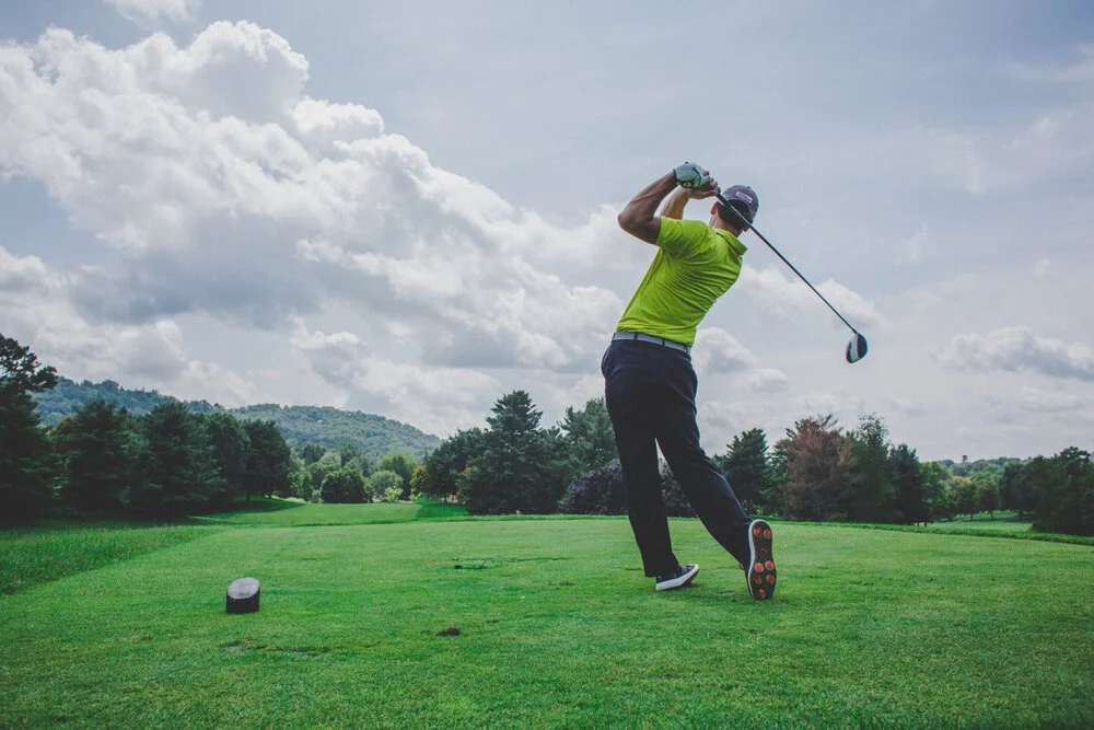 A man in bright green shirt and black pants swinging a golf club on a golf course, with trees and cloudy sky in the background.