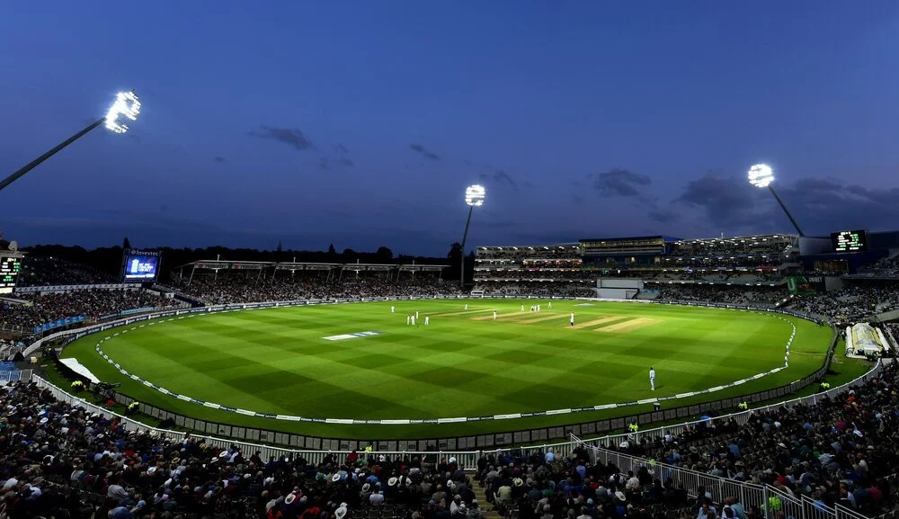 A night-time cricket stadium filled with spectators, illuminated floodlights, and players on the grass field.