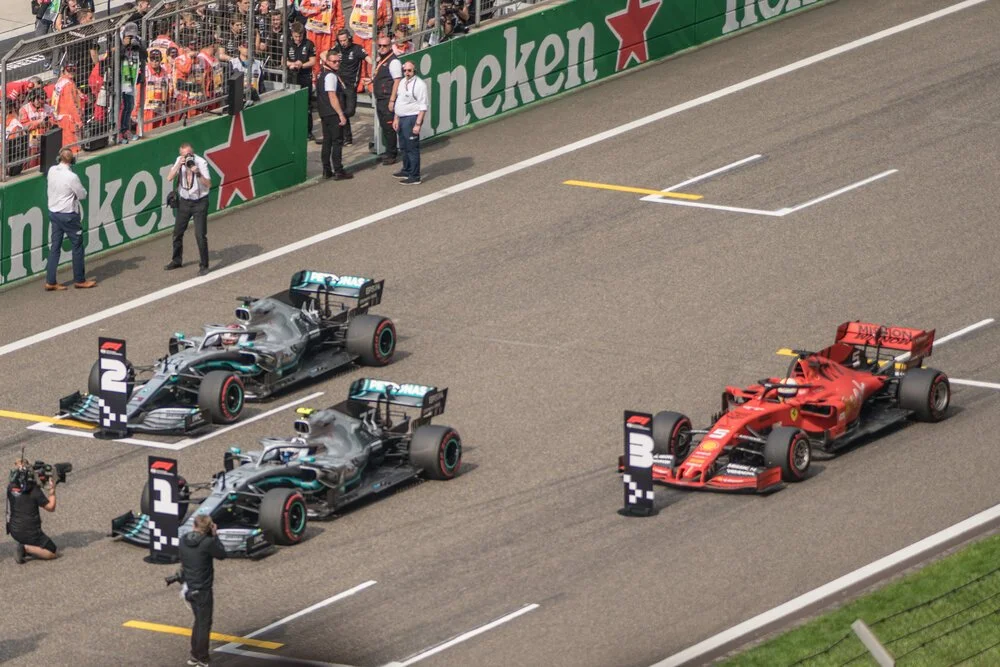 Two Mercedes-AMG Petronas Formula 1 cars and one Ferrari SF-23 Formula 1 car at the finish line during a race, with team personnel and fans in the background.