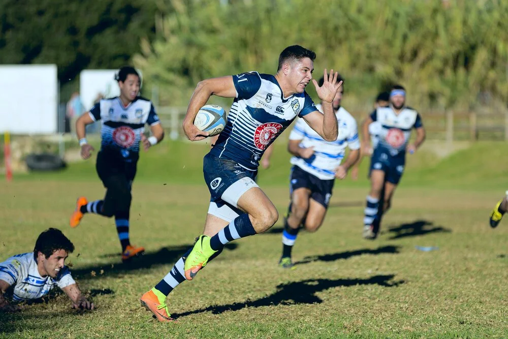 Rugby players in action during a game, with one player sprinting forward holding the ball, while others chase after him on a grassy field.