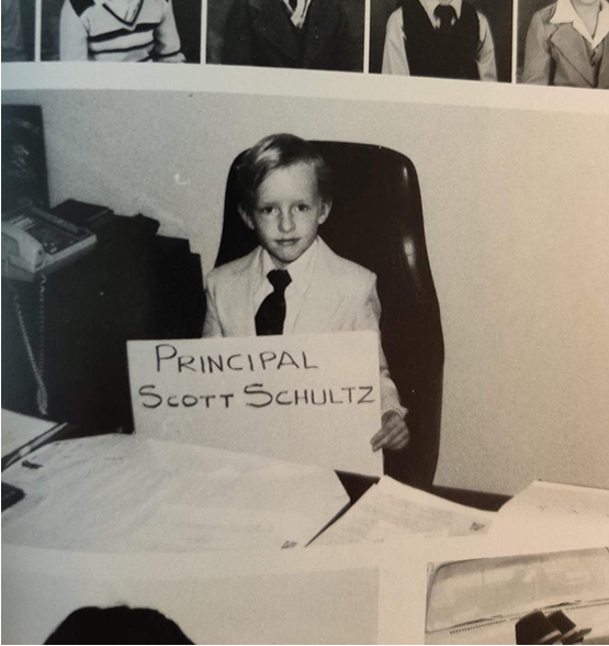 Young Scott Schultz sitting at a desk, holding a sign that reads 'Principal Scott Schultz'.