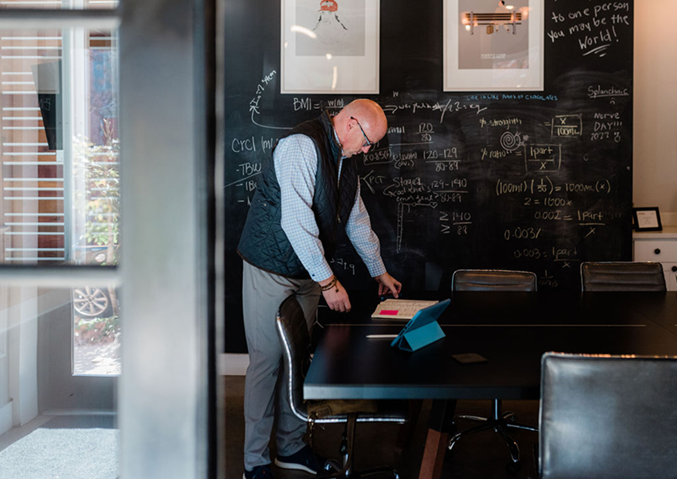 Scott Schultz standing in a conference room with a black chalkboard wall filled with notes and diagrams, working on a laptop and notes.