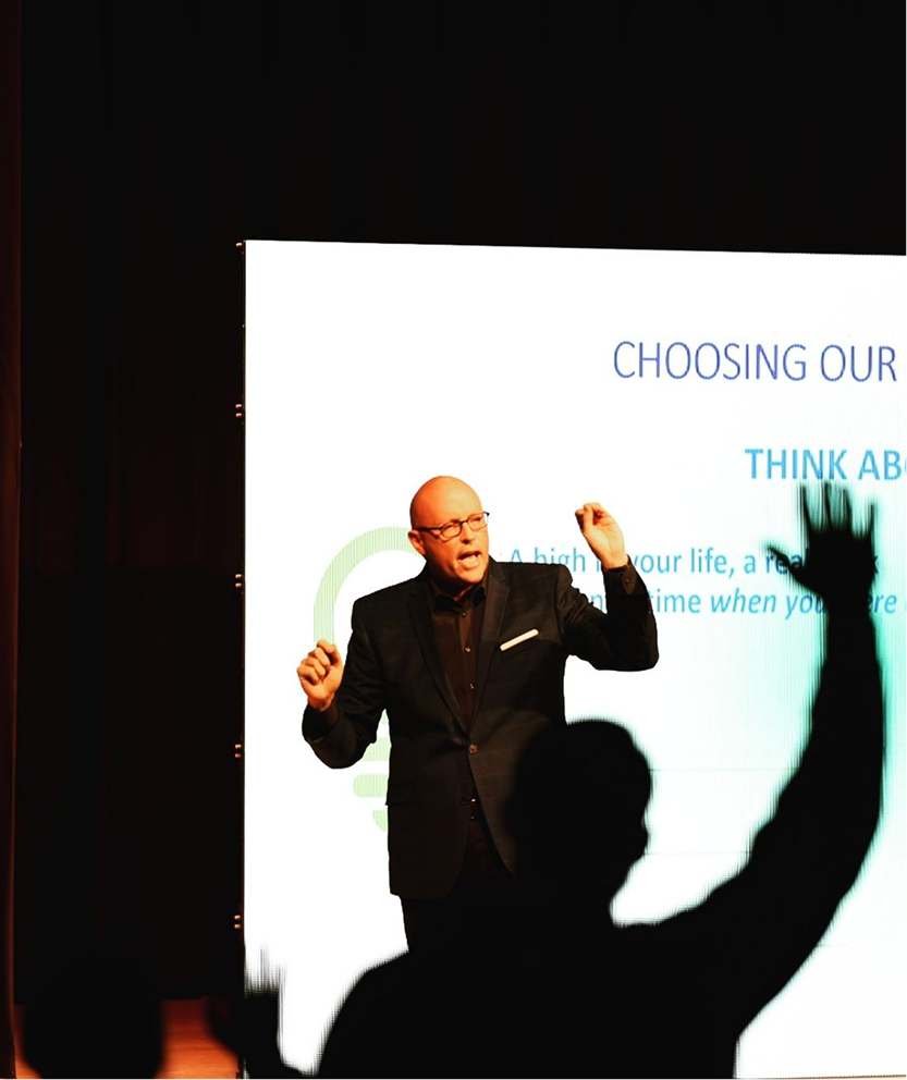Scott Schultz in a black suit speaking at a presentation, with a large screen behind him.