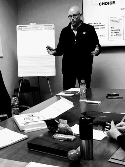 Scott Schultz standing in a conference room holding a phone and gesturing with his other hand, with a flip chart and a poster on the wall behind him.