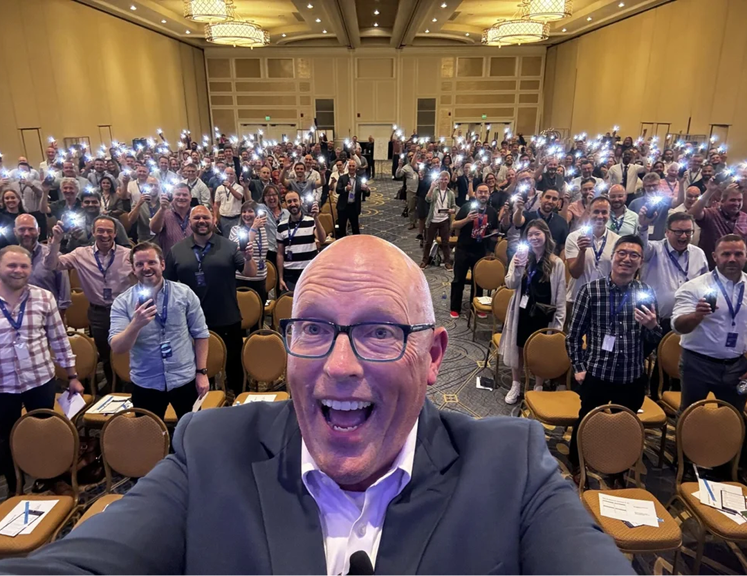 Scott Schultz in a suit with glasses taking a selfie in front of a large audience of people holding up illuminated phones in a conference room.