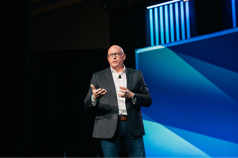 Scott Schultz in a suit giving a presentation on stage with a large screen behind him.