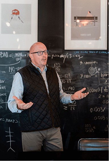 Scott Schultz speaking in front of a blackboard, wearing glasses, a checkered shirt, and a vest, in a boardroom.