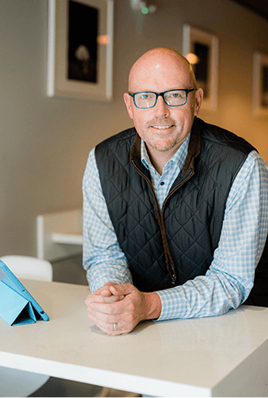 Scott Schultz smiling and leaning on a white table in a warmly lit room.