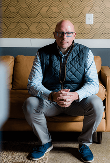 Scott Schultz sits on a couch, with his hands clasped in front of him. He is wearing a blue checkered shirt, a navy quilted vest, gray pants, and blue shoes. The background includes a patterned wall.