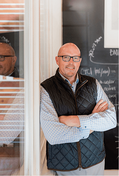Scott Schultz standing with arms crossed and smiling indoors near a black chalkboard.