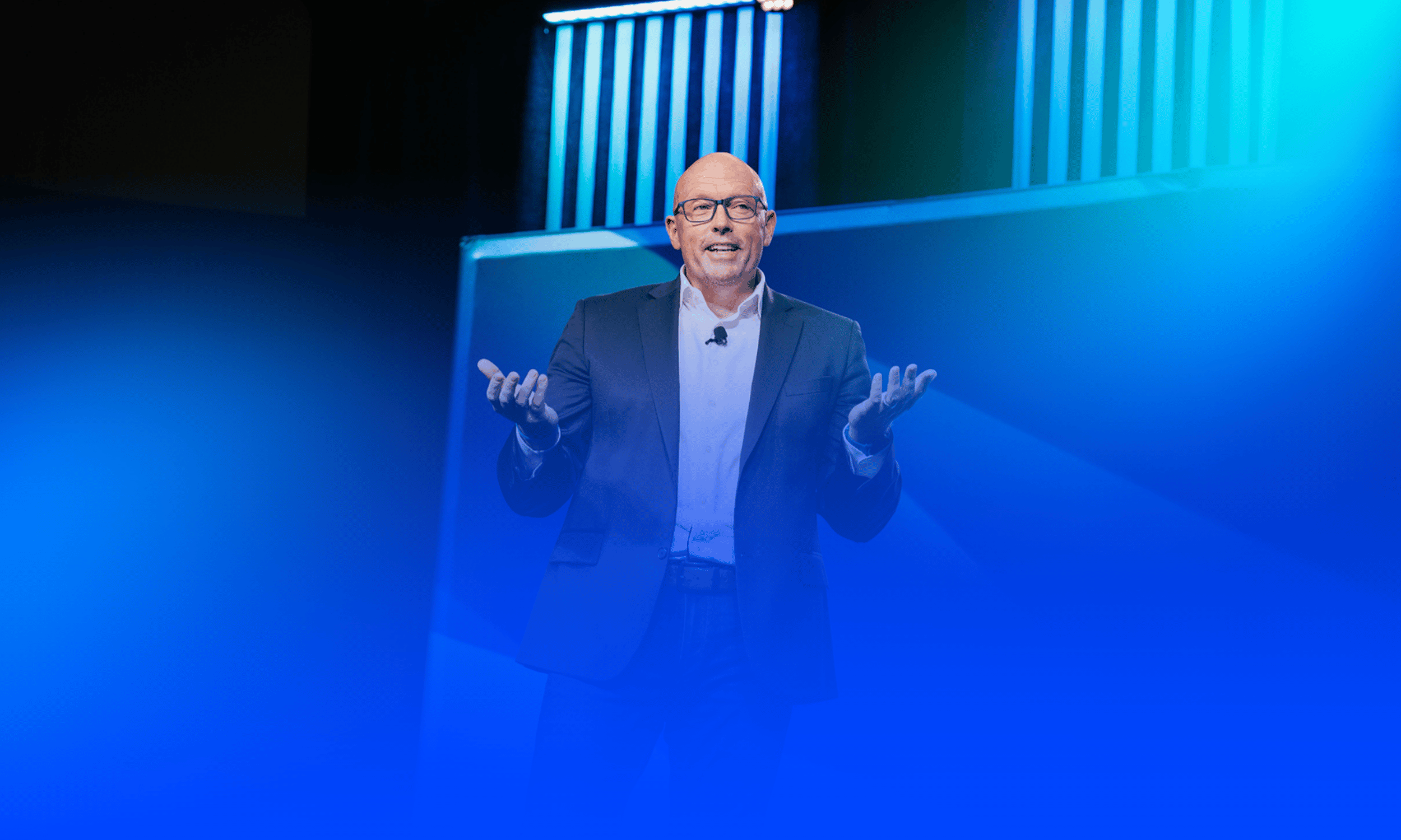Scott Schultz in a suit speaking at a conference, gesturing with his hands, with blue and purple lighting and abstract background structures.