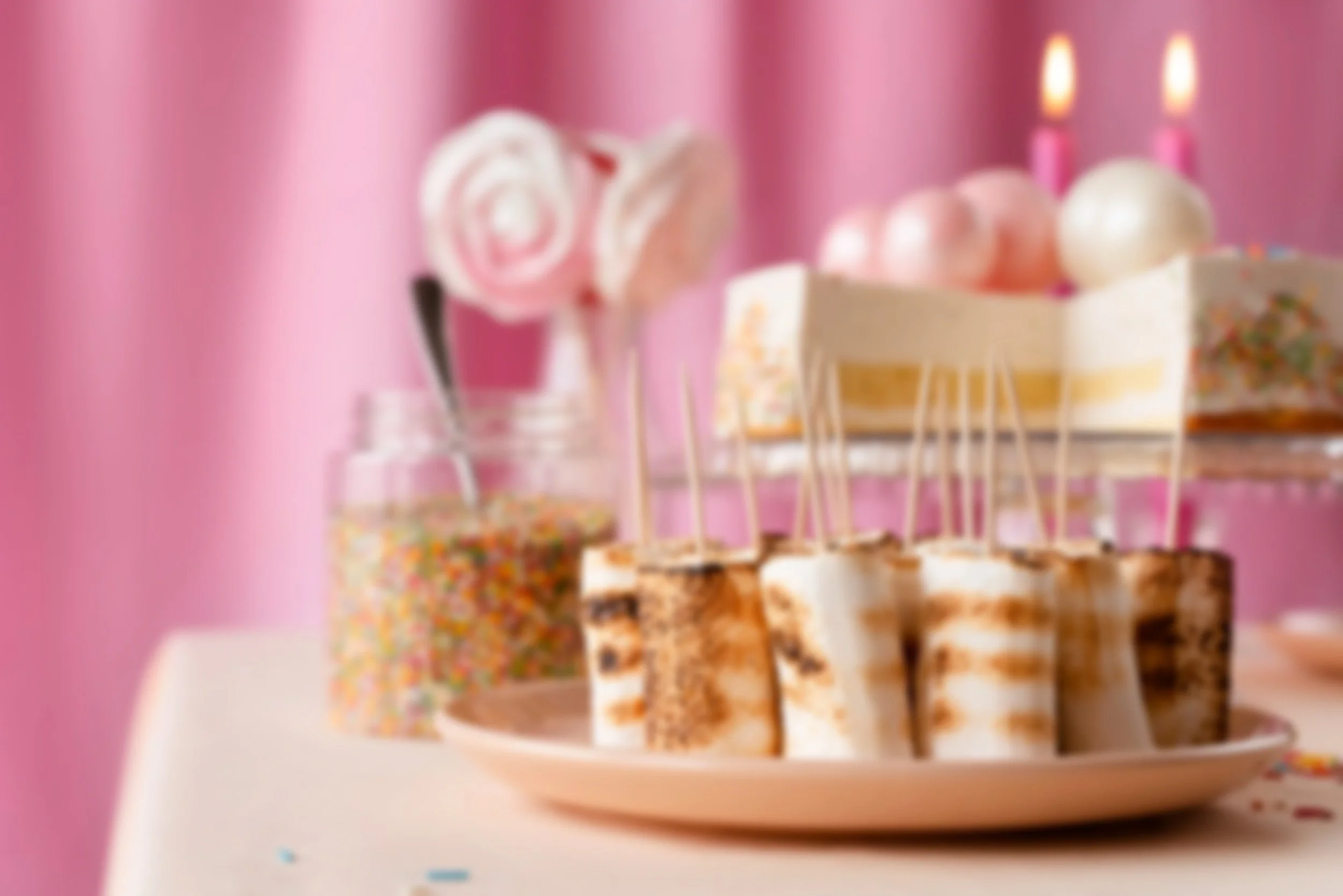 Blurred image of a birthday cake with pink candles, slices of cake on a plate, and a jar of rainbow sprinkles against a pink background.